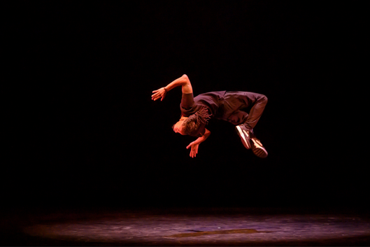 A dancer backflips on a dark stage.